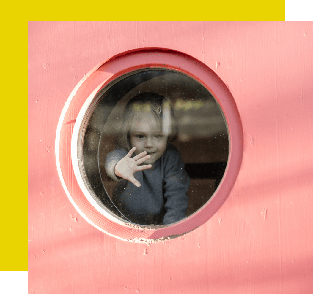Ein Kind schaut durch ein rundes Fenster in einer rosa Wand